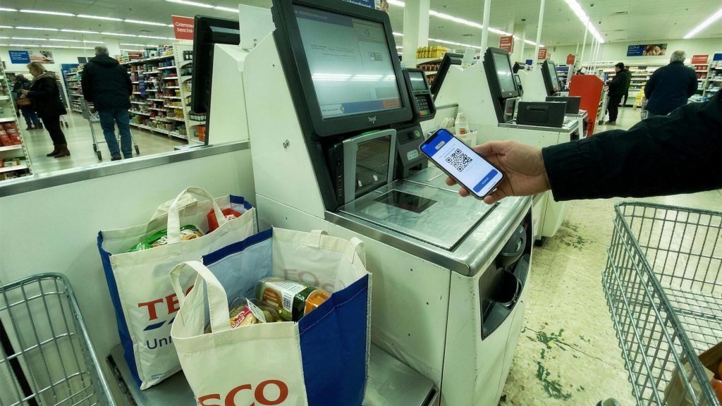 Shopper scanning a digital gift card QR code on a phone at a Tesco self-checkout, illustrating supermarket cashback on a food shop